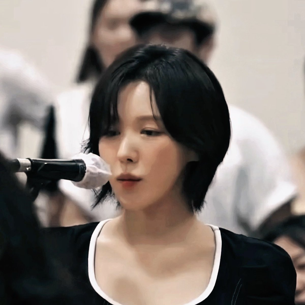 A close-up of a young woman with short black hair looking toward a microphone, wearing a black top with white trim over the neckline.