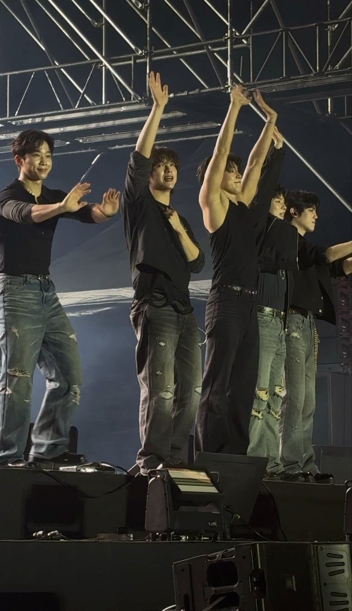 Six members of a K-pop boy group standing on stage in coordinated all-black outfits with distressed jeans, arms raised toward the audience during what appears to be a concert finale.
