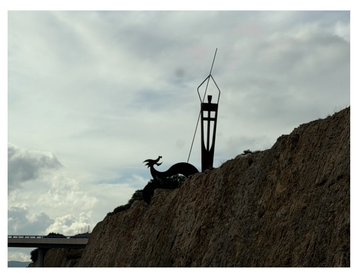 A silhouetted statue of a dragon stands atop a grassy hill beside a tall lamppost against a cloudy sky.