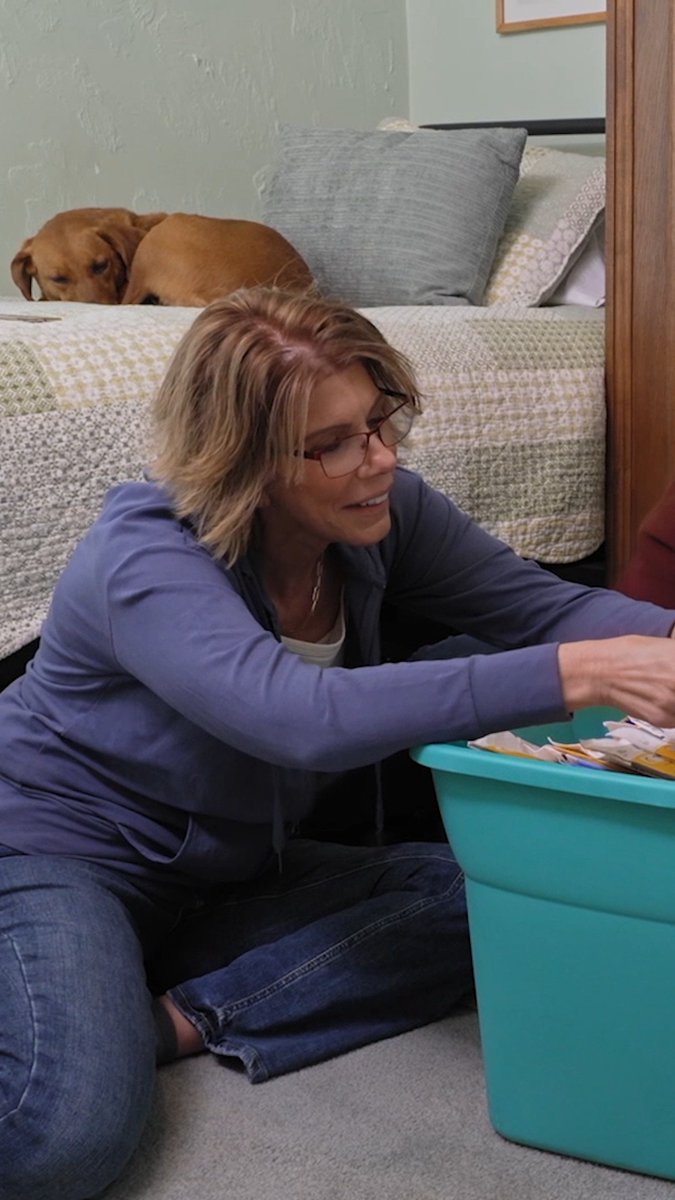 A woman wearing glasses and a purple hoodie kneels on the floor smiling as she sorts through papers in a teal plastic bin, with a brown dog resting on the bed behind her.