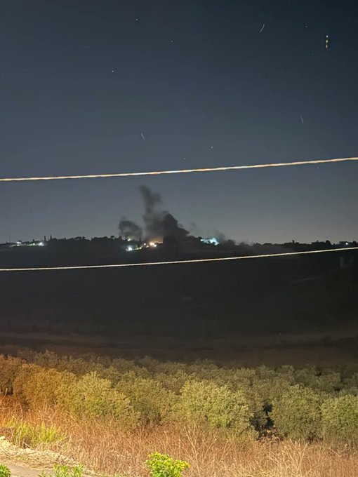 A nighttime view shows distant smoke rising from fires on the horizon, with two illuminated cables crossing the foreground and olive trees visible at the bottom.