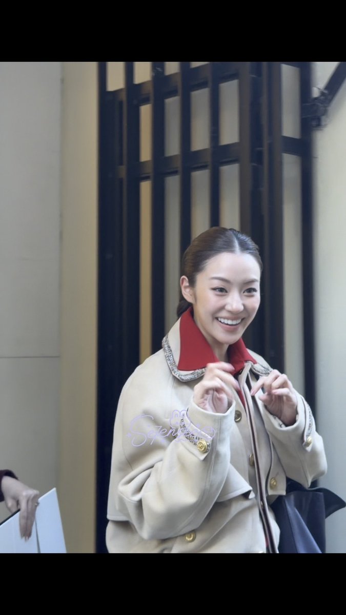 A young woman with her hair pulled back smiles brightly while wearing a beige Chanel coat with a distinctive red collar and gold button details, posing outside what appears to be a Chanel store, fitting the post's tags referencing the PFW Spring/Summer event and LenaMiu.