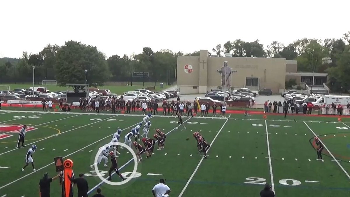 A football game in progress at Archbishop Ryan stadium, with players lined up in formation on the field and a statue of Jesus visible on a building in the background, with a circle highlighting one player.