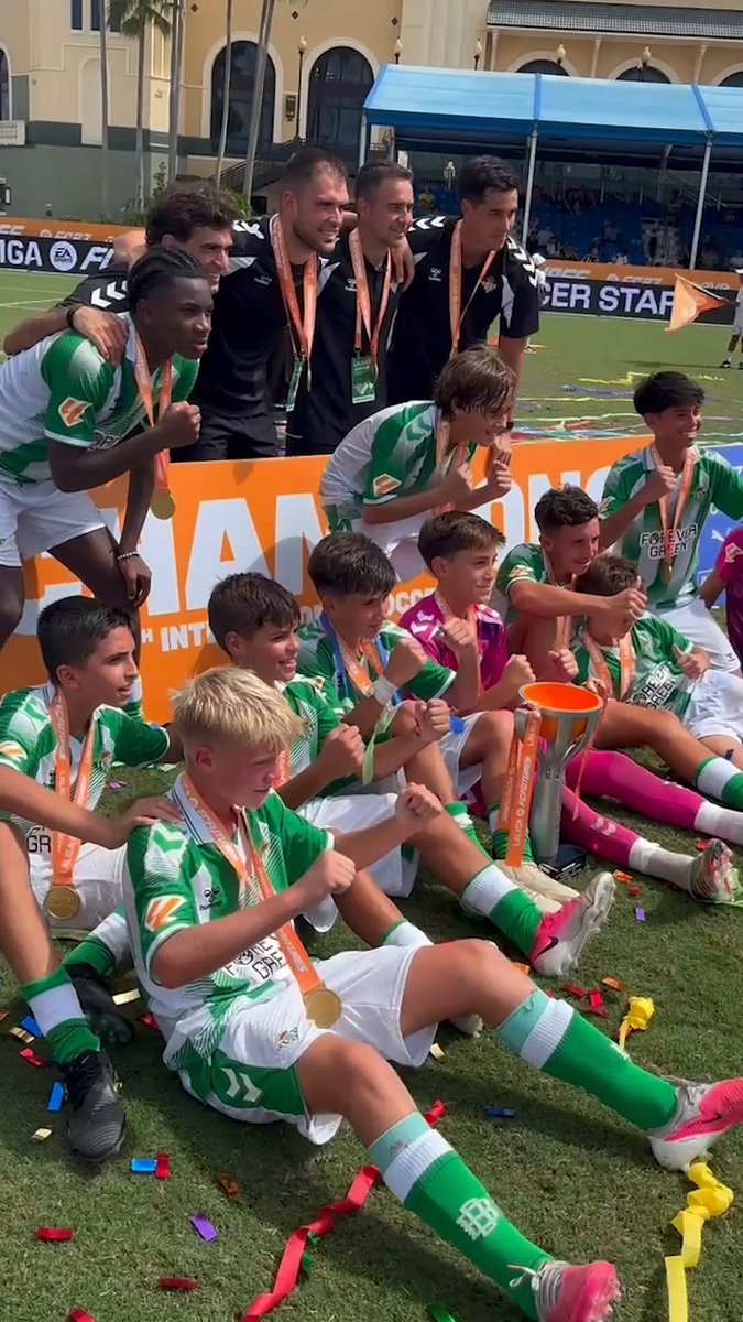 Real Betis youth players and coaching staff celebrate with gold medals and the LaLigaFutures championship trophy on the pitch, wearing their iconic green kits amid scattered confetti following their tournament victory.