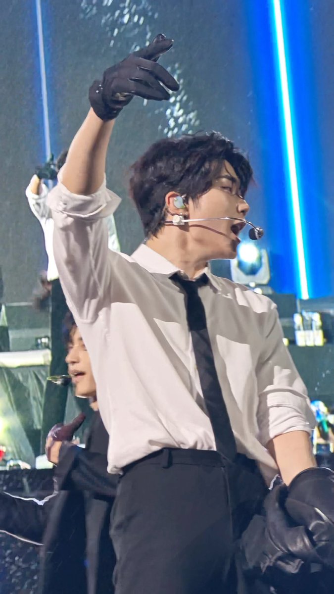 A performer on stage wearing a white dress shirt, black tie, and black gloves, with one arm raised dramatically while singing through a headset microphone.