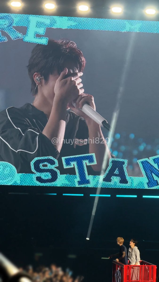A concert photo showing a large screen displaying a performer holding a microphone, with the text 'STAN' visible on screen and a watermark '@inuyasahi820'; two people are visible on stage below the screen.