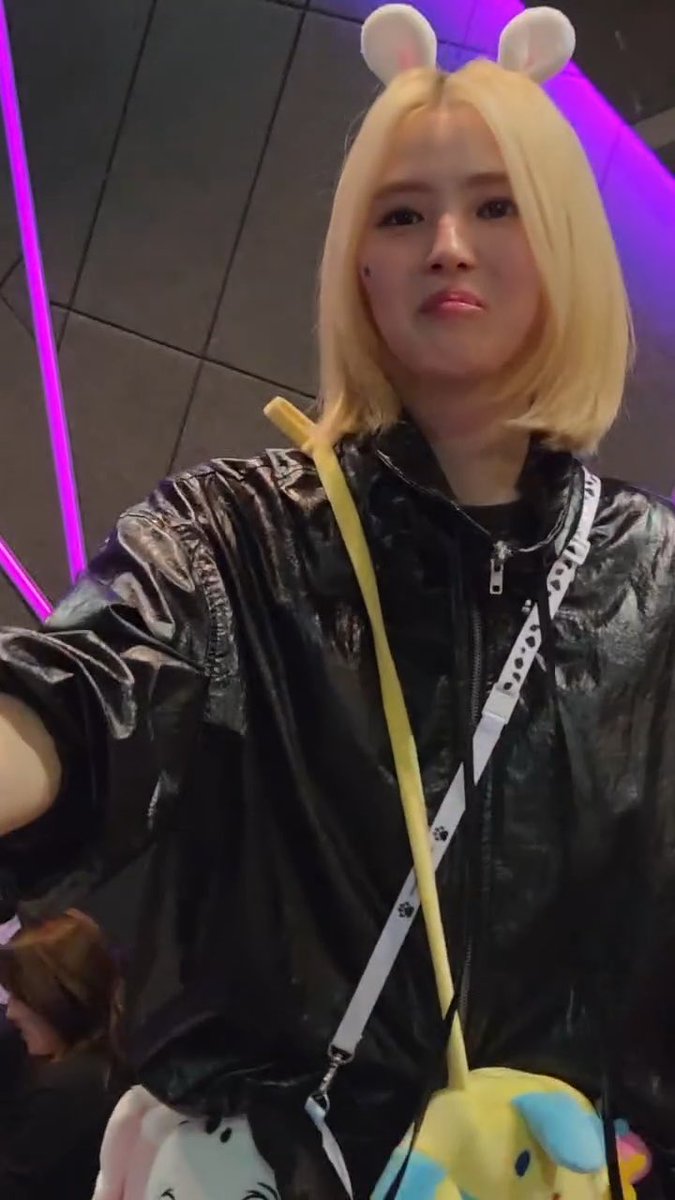 A person with a blonde bob haircut wearing mouse ears and a shiny black jacket, carrying a colorful bag with a white strap in what appears to be an event venue.