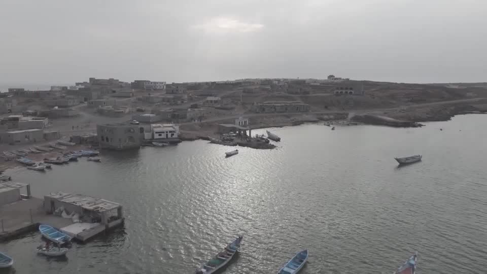 An aerial view of a coastal town with weathered buildings and small fishing boats moored along its shore, evoking a sense of isolation. The image accompanies news of Yemen's leader announcing military action against the Houthis amid ongoing Saudi-backed efforts to counter their advances.