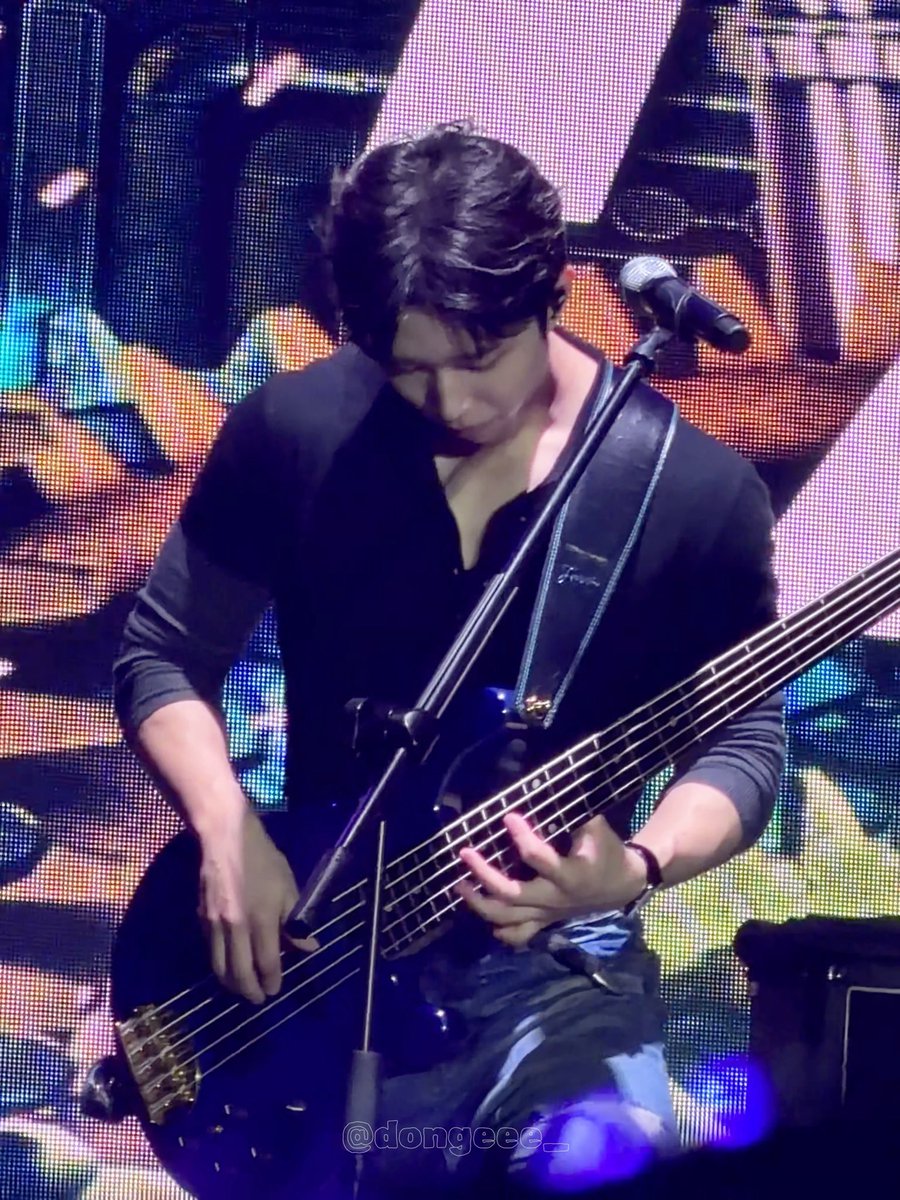 A musician with dark hair plays a bass guitar on stage in front of a microphone, with a colorful LED screen behind him; watermark reads @dongeee_