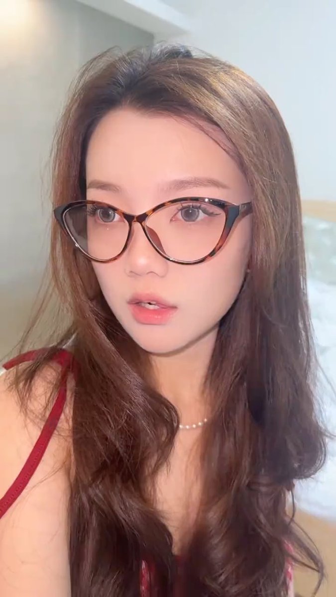 A selfie of a young woman with long brown hair wearing tortoiseshell cat-eye glasses, a delicate pearl necklace, and a red top.