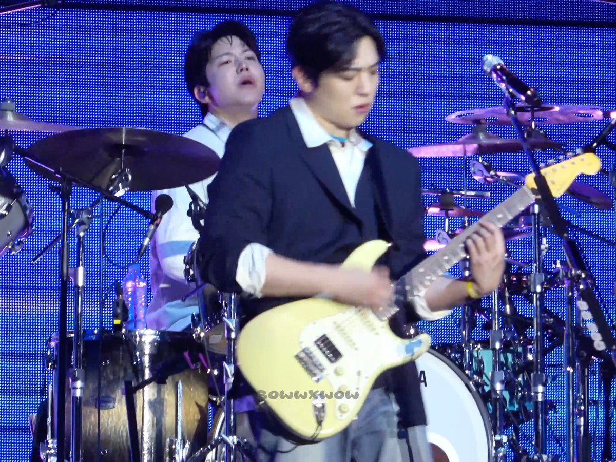 Two male musicians perform on stage against a blue LED background; one plays a yellow electric guitar while the other plays drums, with a 'BOWWOW' watermark visible.