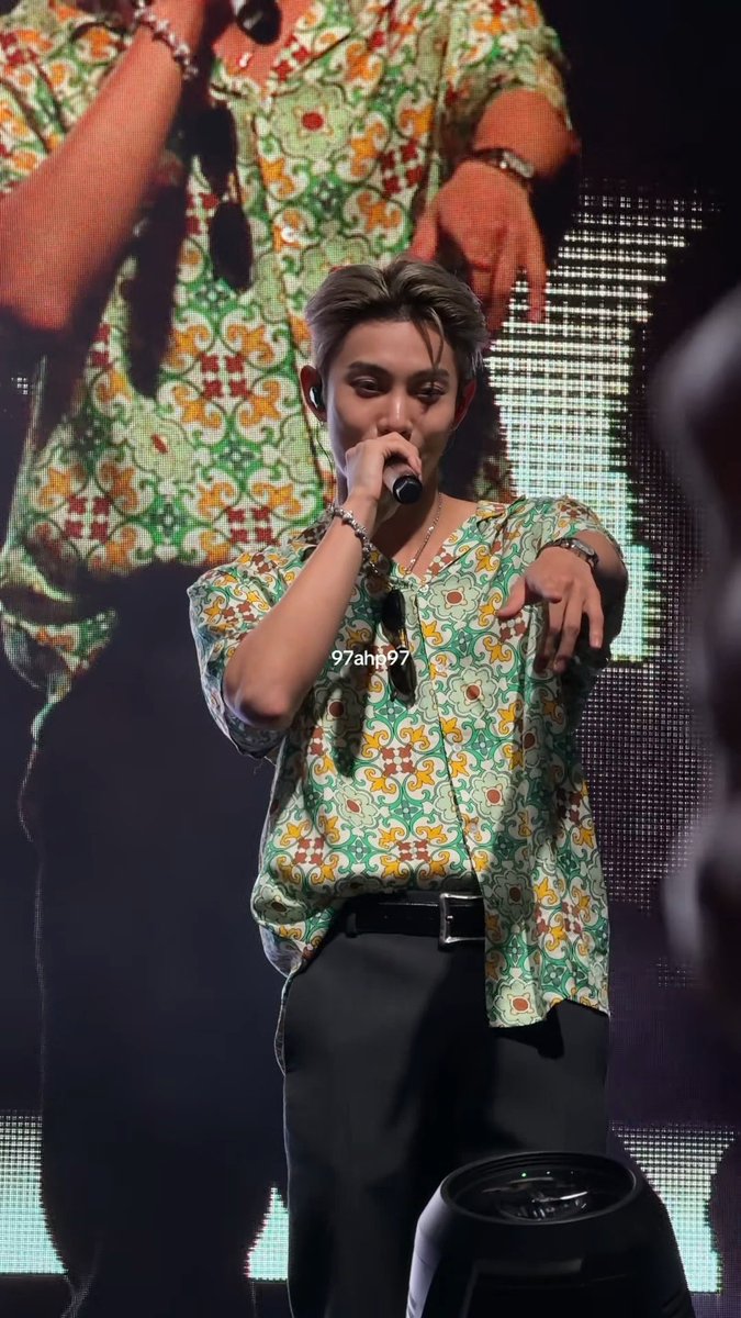 A performer on stage holding a microphone, wearing a green and white patterned shirt, with the text '97ahg97' superimposed on his chest.