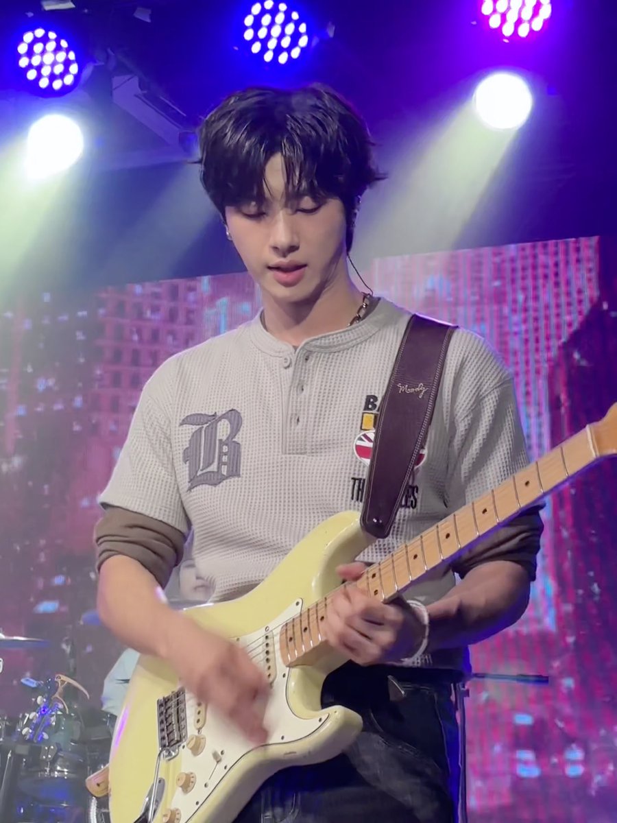 A young male musician with dark hair plays a cream-colored electric guitar on a stage lit by purple and blue lights, wearing a white jersey with 'B' and 'THE' visible.