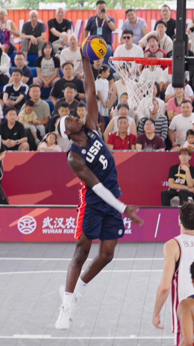 A basketball player wearing a dark blue USA jersey with the number 6 leaps toward the hoop for a dunk during a 3x3 basketball game in Wuhan, with a 'WUHAN RURAL' banner and spectators visible.
