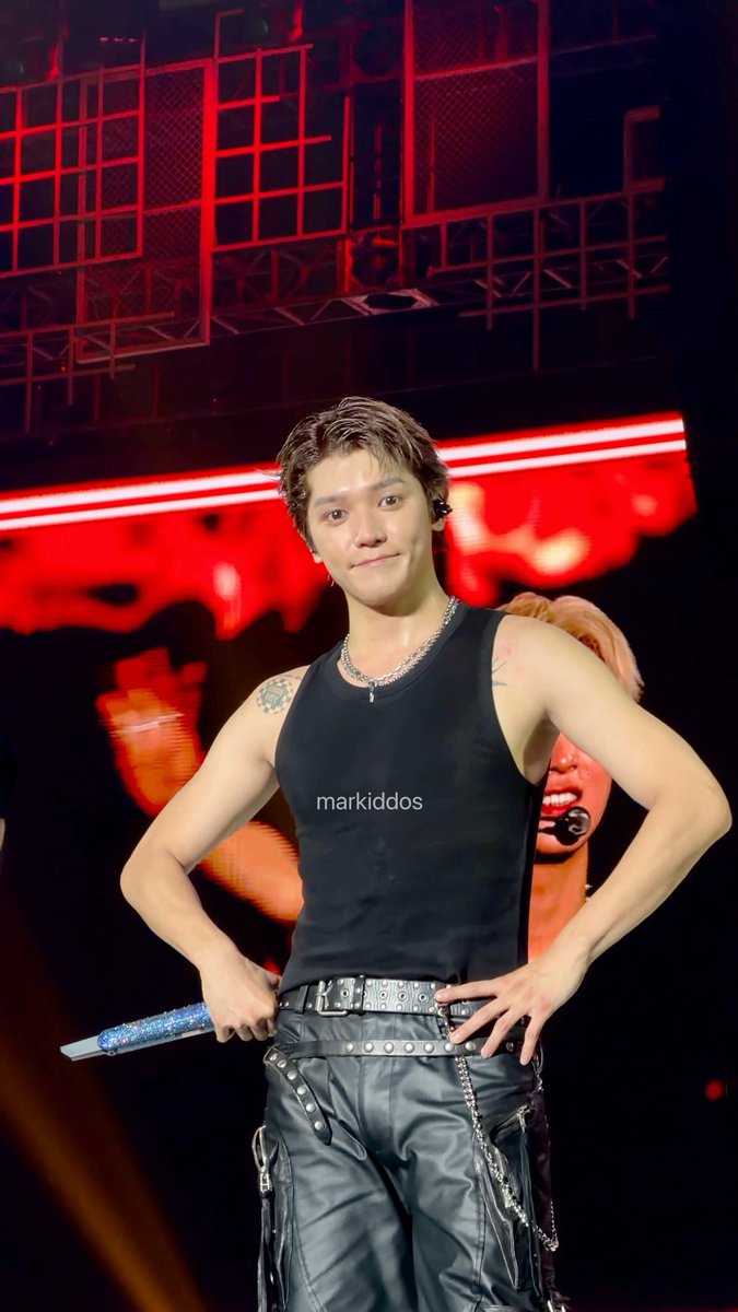 A young man with curly hair in a black tank top and leather pants poses on stage holding a glittery blue microphone, with a watermark 'markiddos' visible.