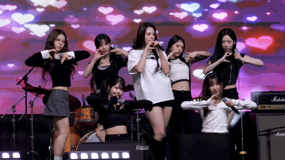 Seven female performers on stage making heart gestures with their hands, holding them against a backdrop of pink hearts displayed on screens. The group is performing with microphones, and Marshall amplifiers are visible on stage.