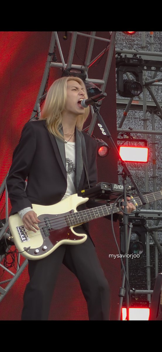Blonde performer in a black blazer singing and playing a cream-colored bass guitar on stage with red lighting, watermarked 'mysaviorjoo'.