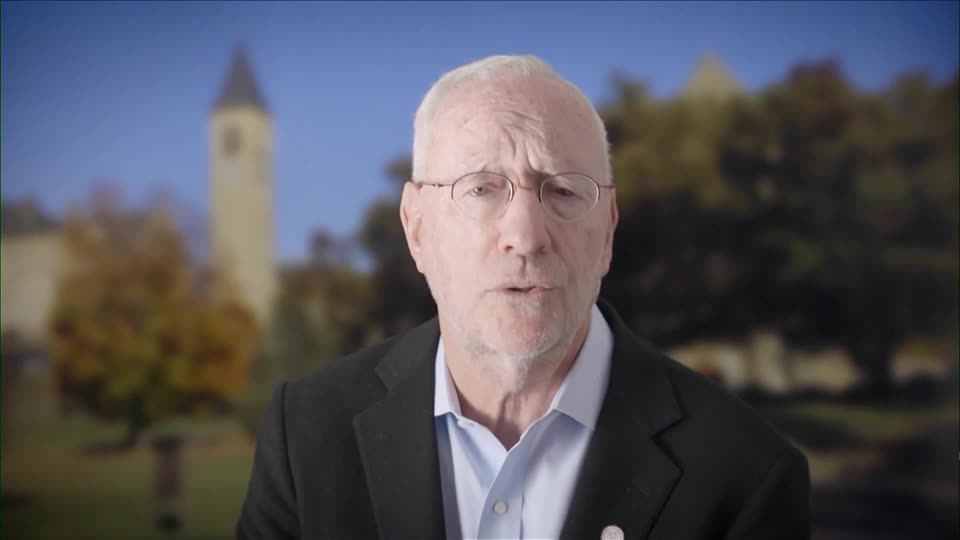 An older man with glasses and a gray beard, wearing a dark blazer and light blue shirt, speaks to the camera with a university campus featuring a clock tower visible in the blurred background. The image accompanies an article about Cornell University President Michael Kotlikoff addressing fraternity and sorority policies following rape allegations against Chi Phi fraternity members.