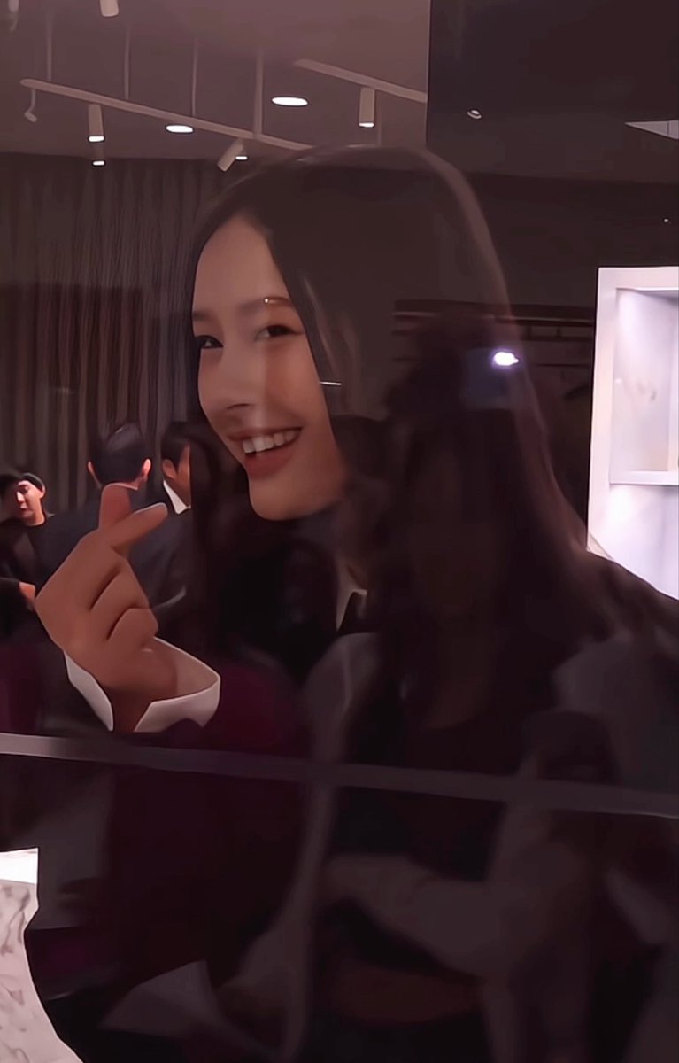 A selfie of a smiling young woman with long dark hair making a finger heart gesture, taken through glass at what appears to be an evening event with other people visible in the background.