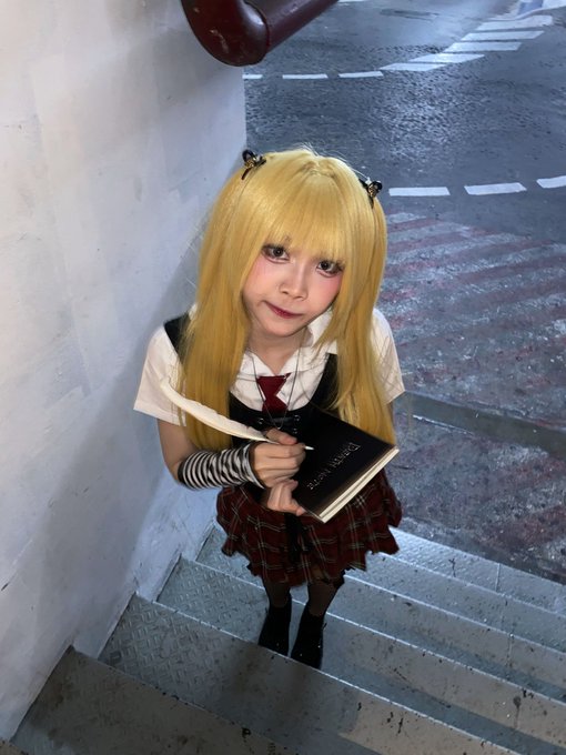 A young person with long blonde hair cosplays as a character, wearing a school uniform with a red tie and plaid skirt, holding a book on a stairwell.