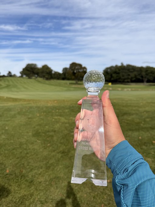 A hand holds a crystal "Member-Member Champions Gross Winner" trophy featuring an etched golf ball design, set against a scenic golf course backdrop. The text on the trophy confirms the victory referenced in the post, celebrating an overall member-member championship win.