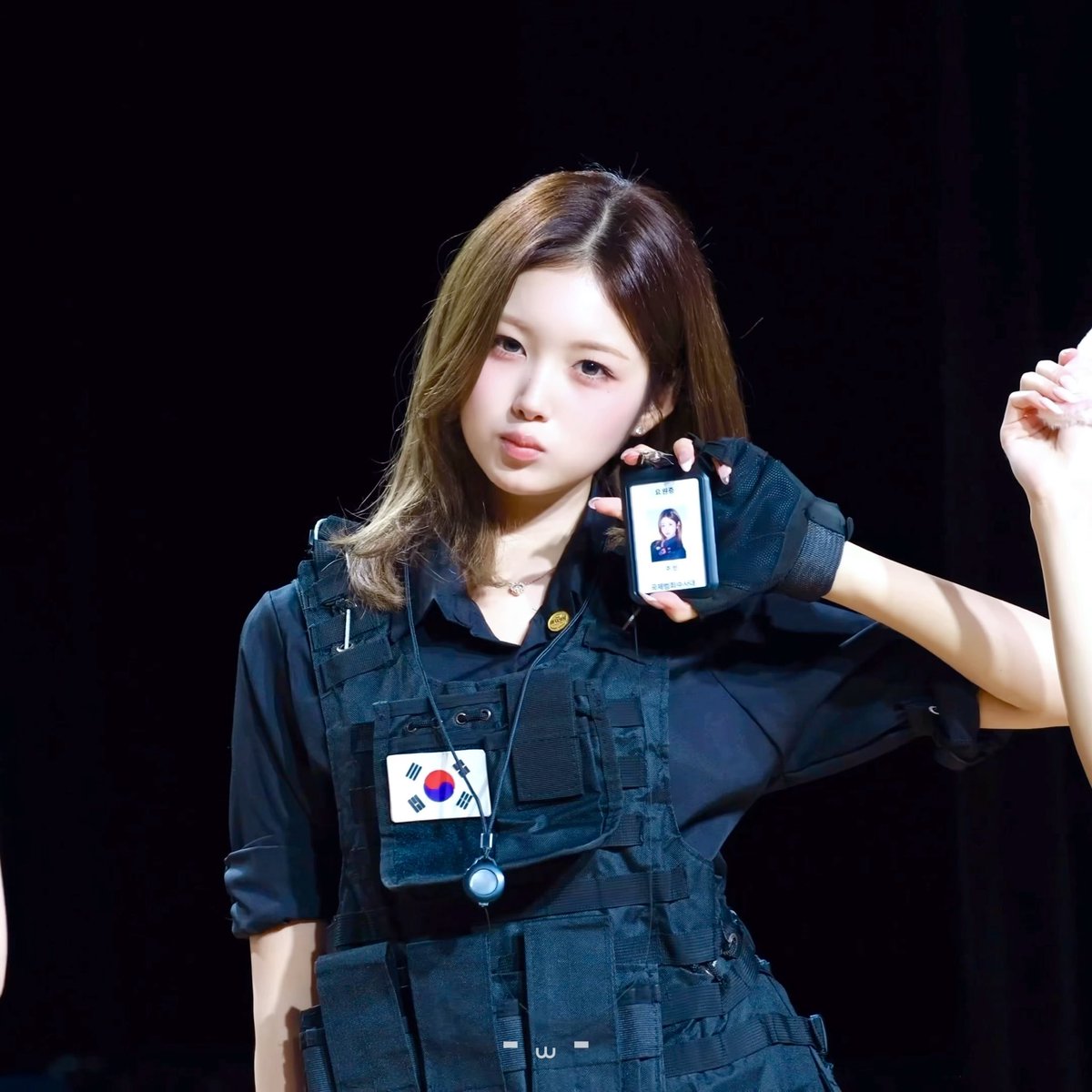 A young woman with light brown hair wears a black tactical vest with a South Korean flag patch, displaying a digital ID badge while posing on a dark stage.