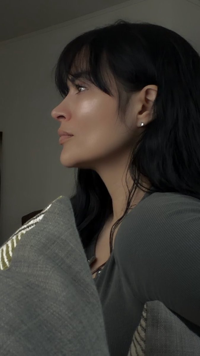 A side profile portrait of a young woman with long black hair, wearing a gray ribbed top and small earrings, in a dimly lit room.
