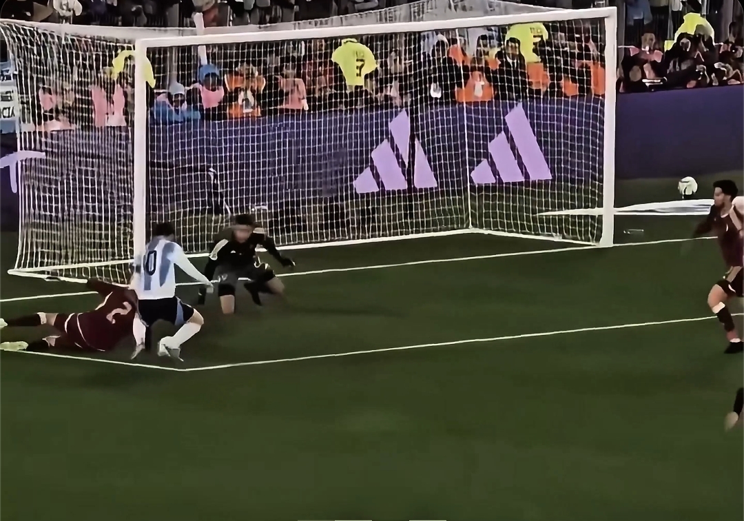 A still from a soccer video game showing a player scoring a goal past the goalkeeper into a net branded with the Adidas logo, while a crowd watches in the background.