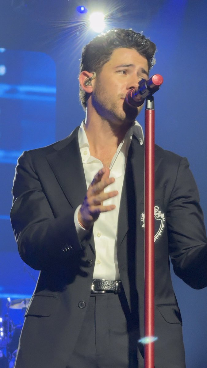 A man, appearing to be Nick Jonas, performs on stage singing into a microphone, dressed in a dark suit with a white shirt, illuminated against a blue-lit backdrop. The image connects to the post's playful caption about his on-stage presence and gesture, showing him mid-performance with an expressive hand movement.