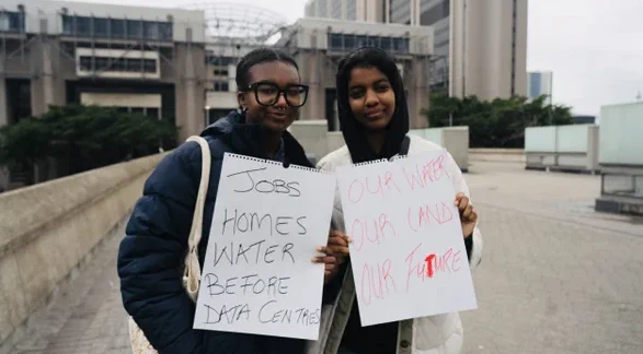Two individuals are holding handwritten signs in an urban setting. One sign reads, "Jobs, Homes, Water Before Data Centres," and the other reads, "Our Water, Our Land, Our Future." The background features modern architectural structures and greenery.