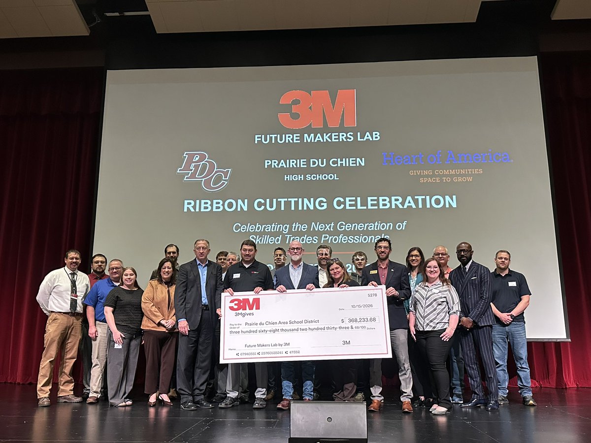 A large group of people stand on a stage behind an oversized ceremonial check, with a projected event announcement and organization logos on the screen behind them. Red curtains frame the stage. Text appears throughout the image.