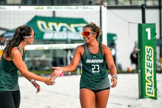 Two volleyball players from the UAB Blazers are on a sand court, smiling and exchanging a handshake. The net and a post with the number 1 are visible in the background. Both are wearing their team uniforms with "Blazers" written on them.
