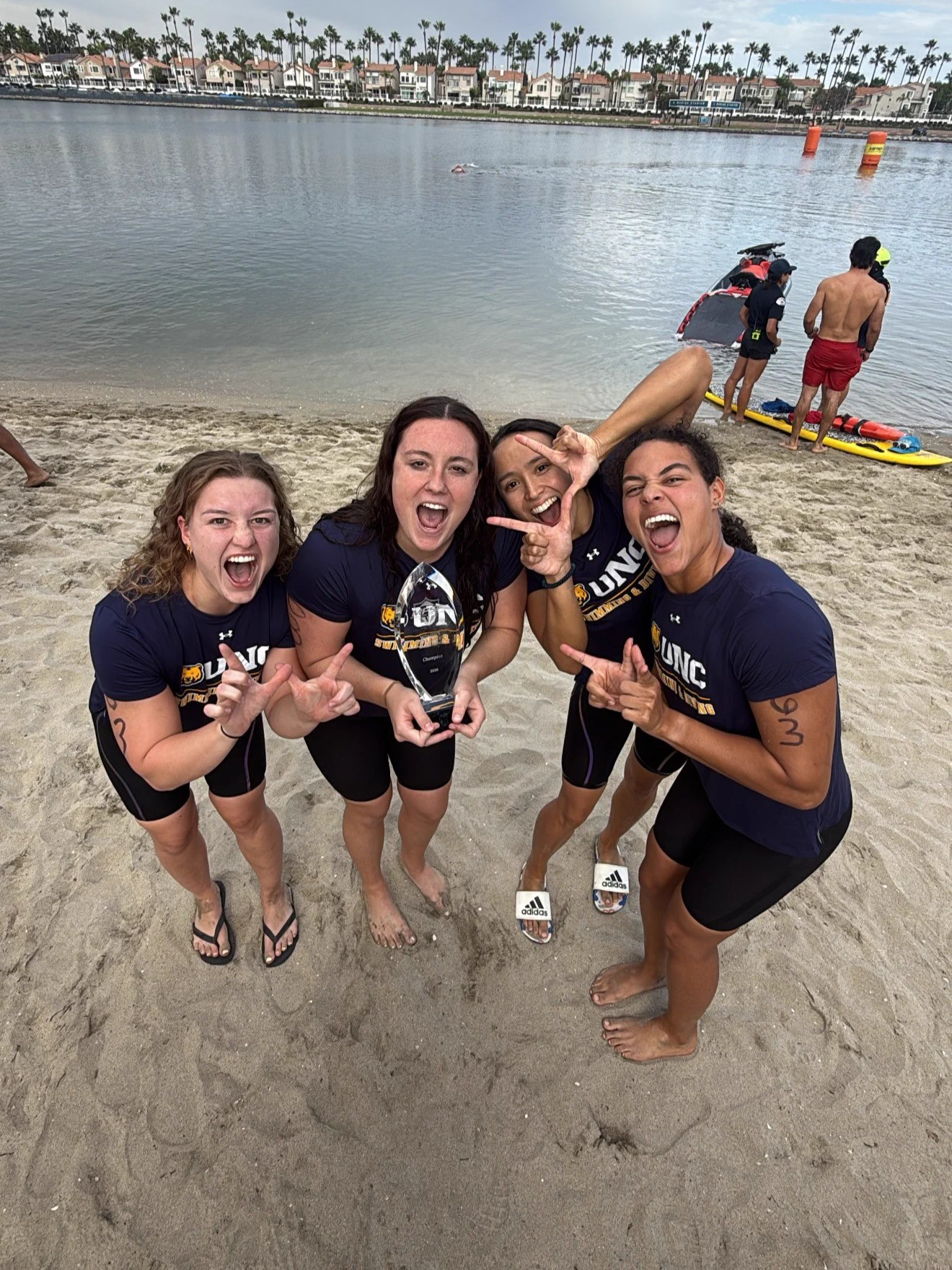 Four people stand on a sandy beach, smiling and cheering while holding a trophy. They are wearing athletic gear with "UNC" printed on it. In the background, there's a body of water with paddleboarders and buildings lined with palm trees. The sky is partly cloudy.