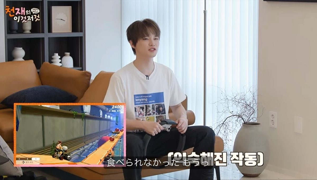 A screenshot from a Korean variety show featuring a young man in a white graphic t-shirt sitting on an orange couch holding a controller, with an inset gameplay screen of Mario Kart showing a score of 2400 and Korean subtitles.
