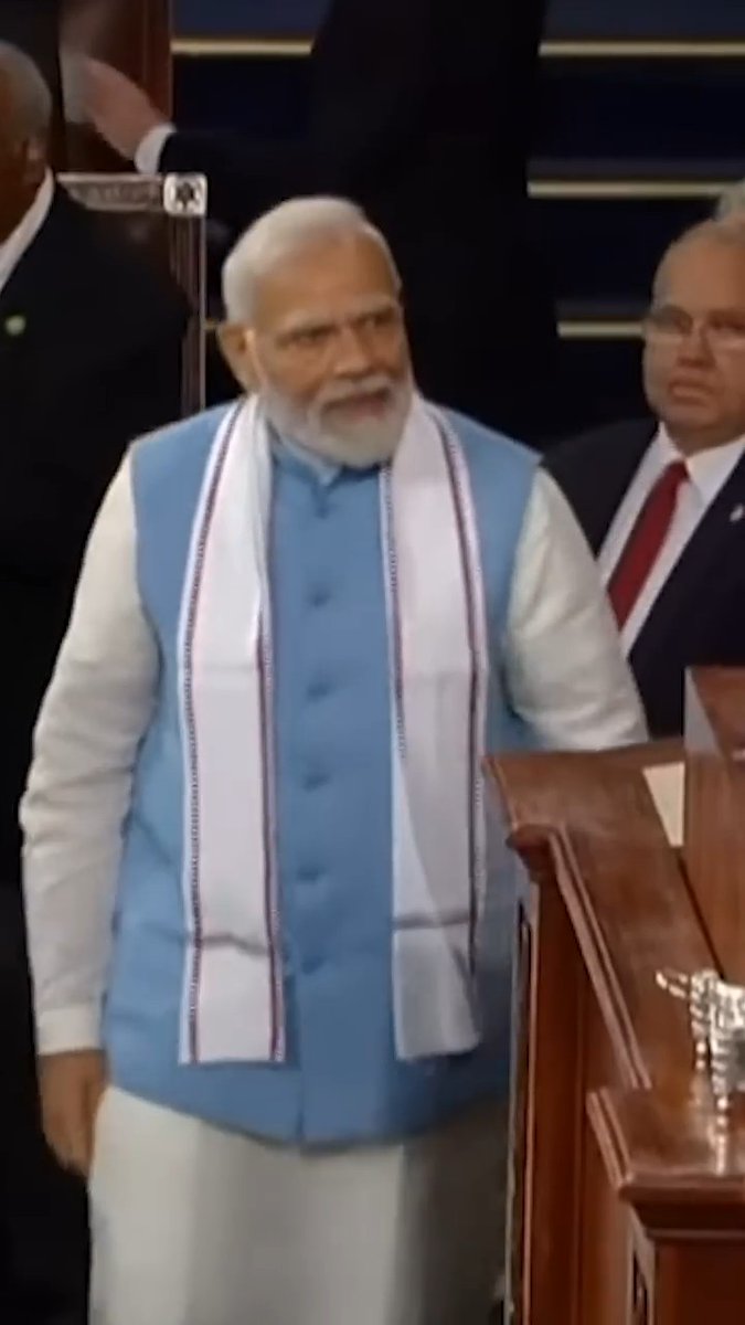 Narendra Modi, the Prime Minister of India, stands at a wooden podium wearing a light blue vest and a white scarf, with another man in a suit visible behind him.