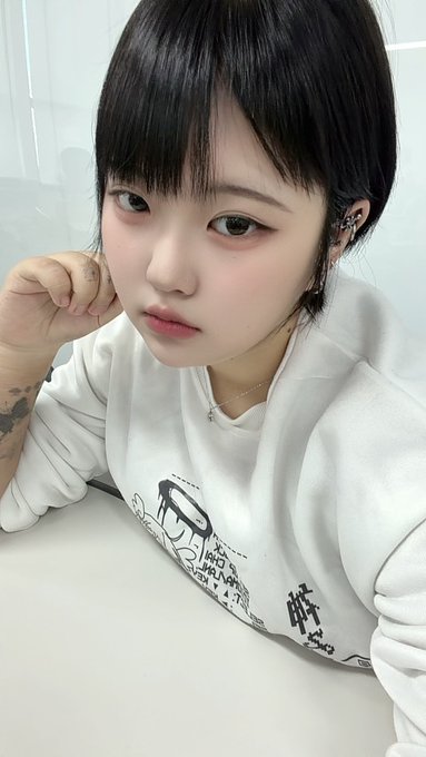 Selfie of a young woman with short dark hair wearing a white sweatshirt with graphic prints, posing with her hand near her face.