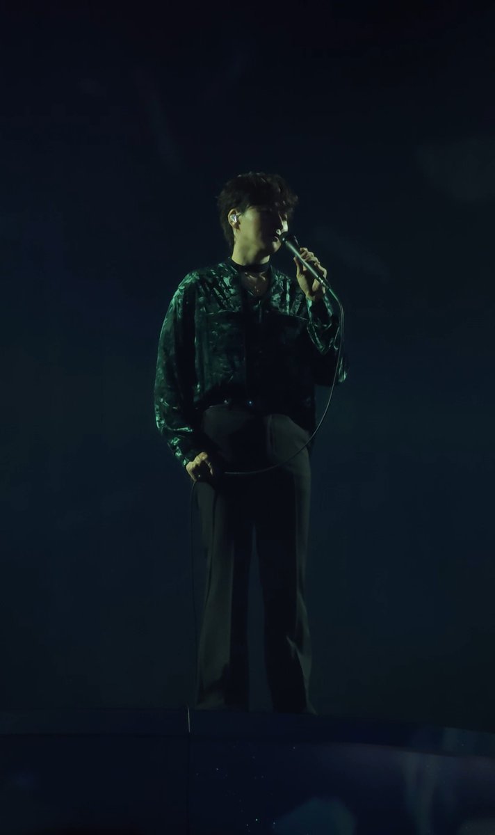 A male performer in a green velvet shirt sings into a microphone on a dimly lit stage during a concert.