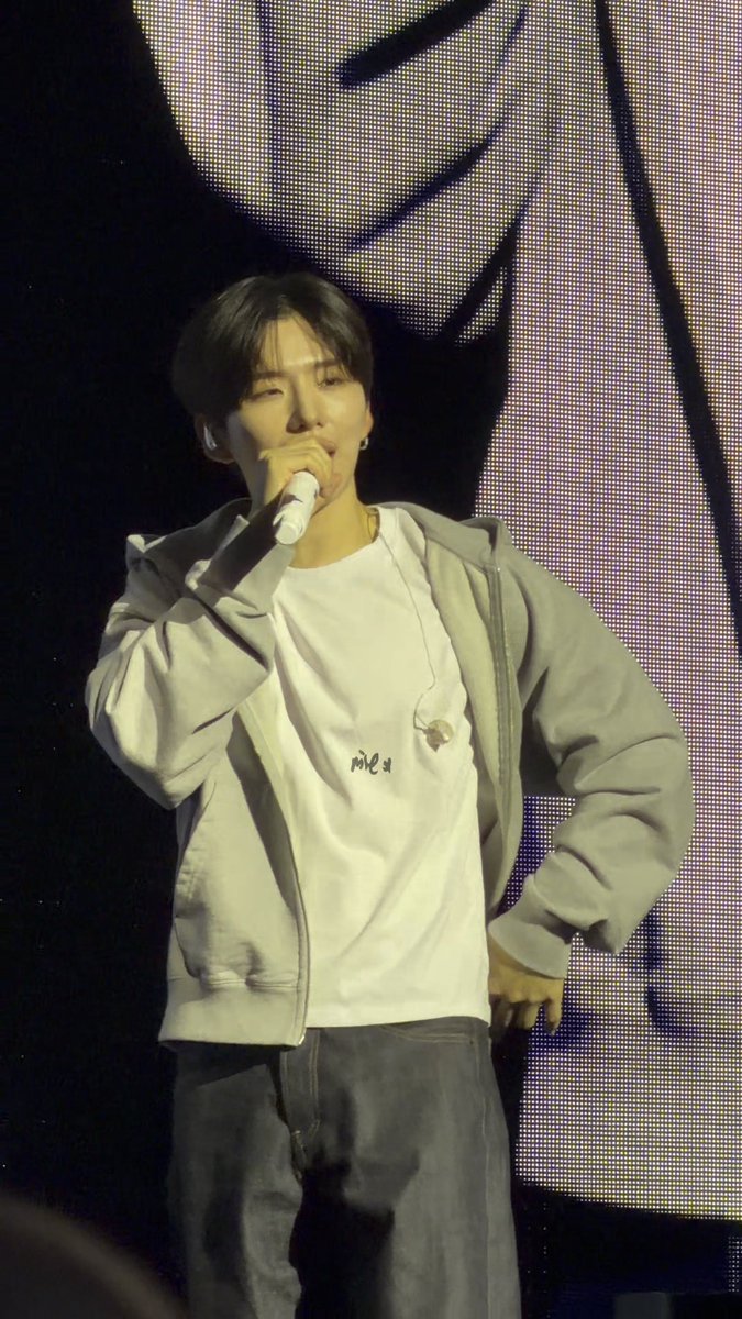 A male performer on stage holding a microphone, wearing a white t-shirt under a light gray zip-up jacket and dark jeans, standing before a large digital screen.
