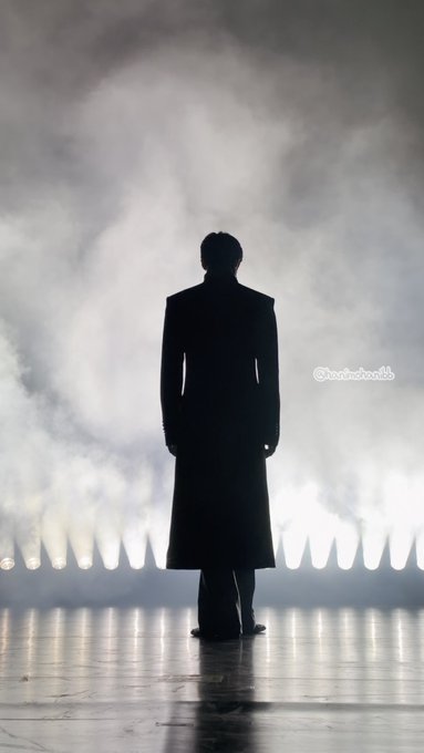 A silhouette of a person in a long coat stands facing away from the camera amid bright white stage lights and smoke, with text '@andrewkho_' visible.