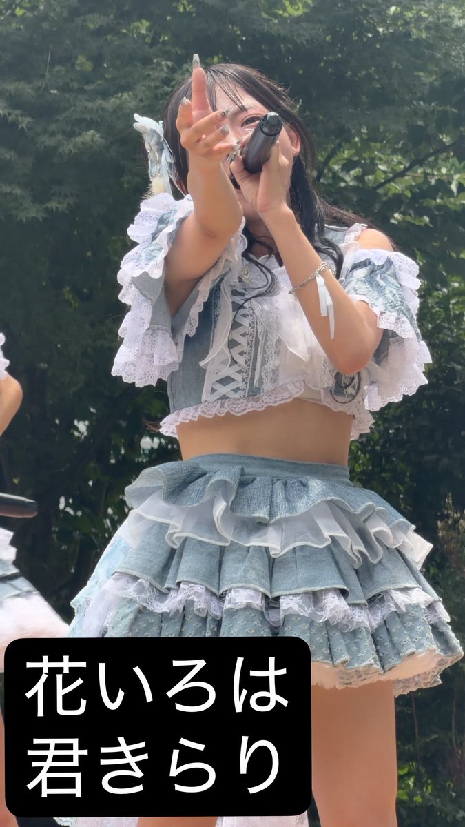 A young woman in an elaborate light blue and white ruffled outfit performs with a microphone outdoors, with the Japanese text '花いろは 君きらり' displayed in a black box at the bottom.