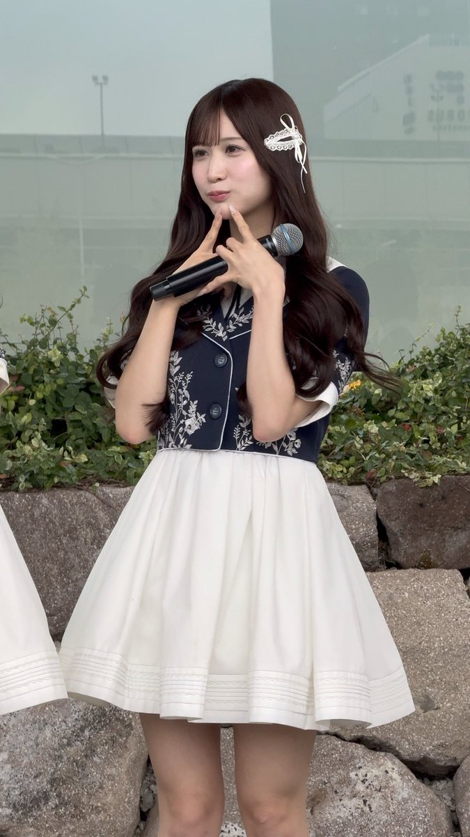 A young woman wearing a navy and white dress with a floral hair accessory holds a microphone while posing outdoors.