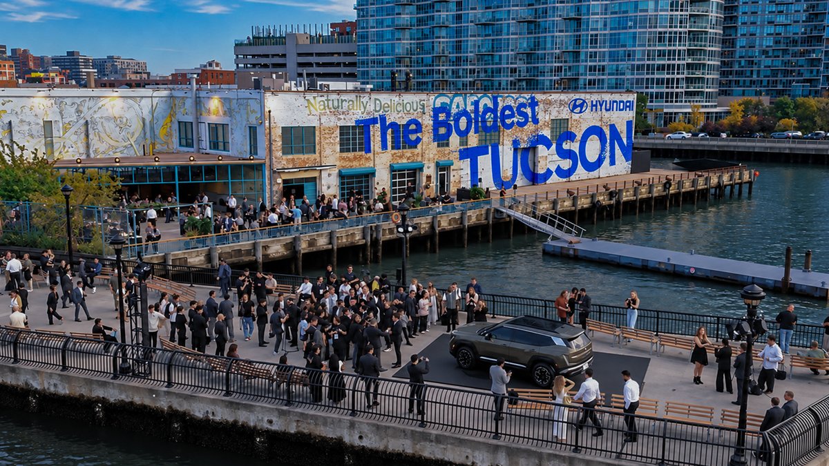 An aerial view of a waterfront promotional event for the Hyundai Tucson with large blue text on a brick building reading 'The Boldest TUCSON', showing a crowd gathered around the displayed vehicle.