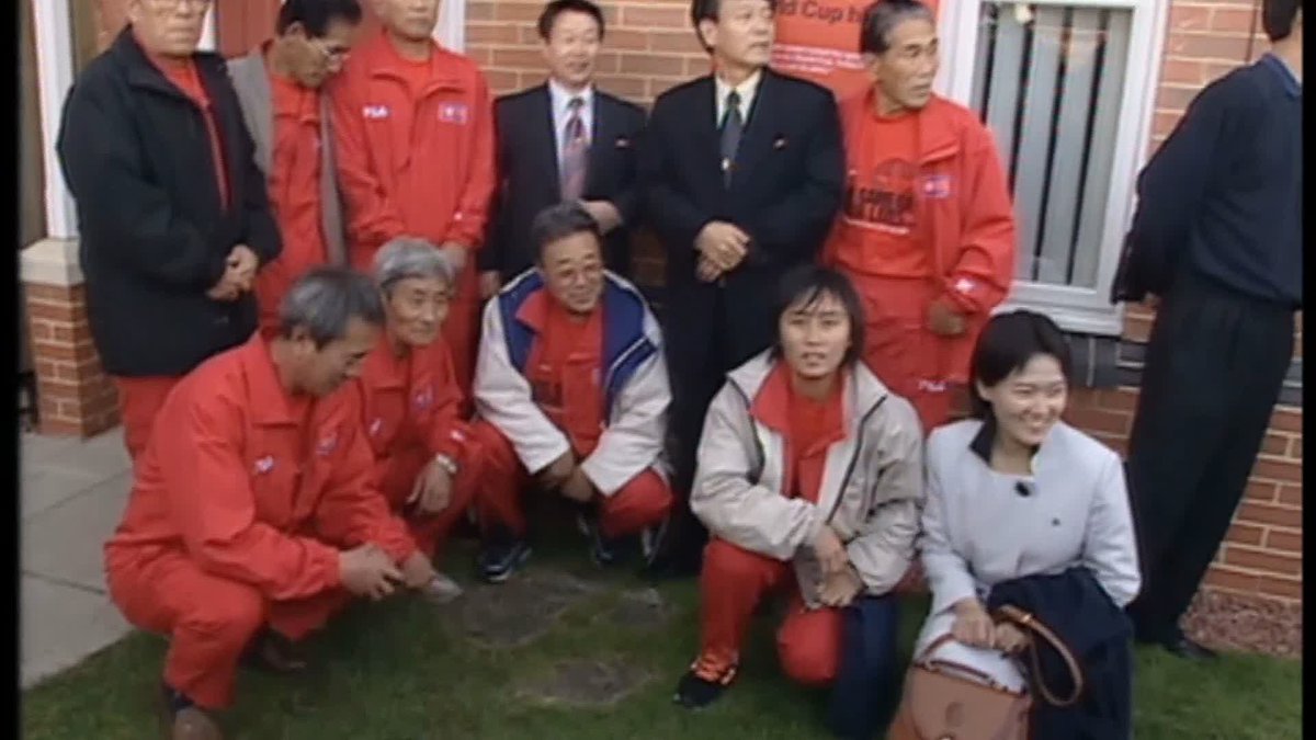 A group photo showing the 1966 North Korean World Cup team, dressed in red FILA tracksuits, posed together with a young North Korean woman, apparently during their return visit to Middlesbrough, England, which is referenced by the partial "World Cup" text on a sign in the background.