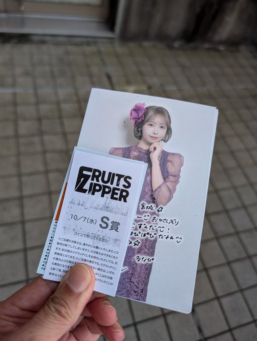 A hand holds a Fruits Zipper signed CD and an event ticket dated '10/7(水)' with 'S賞' visible, featuring a member with short hair wearing a purple outfit.