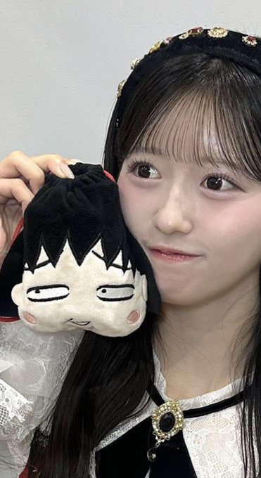 A young woman with long dark hair and bangs, wearing a dark velvet headband with jewels, holds a chibi-style plush toy of a tired-looking character with dark hair and red cheeks next to her face.
