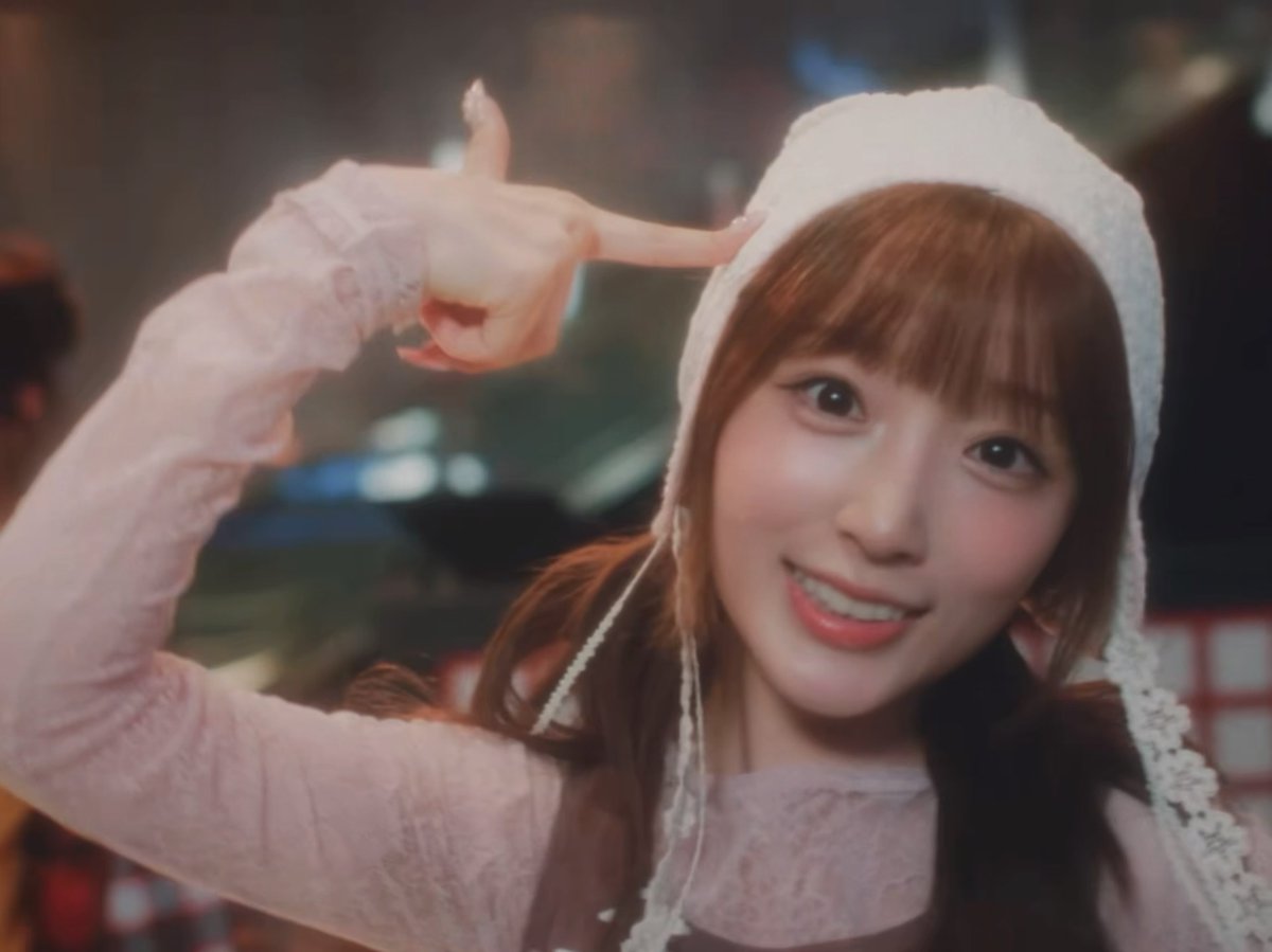 A young woman with light brown hair and bangs wears a white knit bonnet and pink lace top, smiling brightly while pointing her finger at her head in a playful gesture.