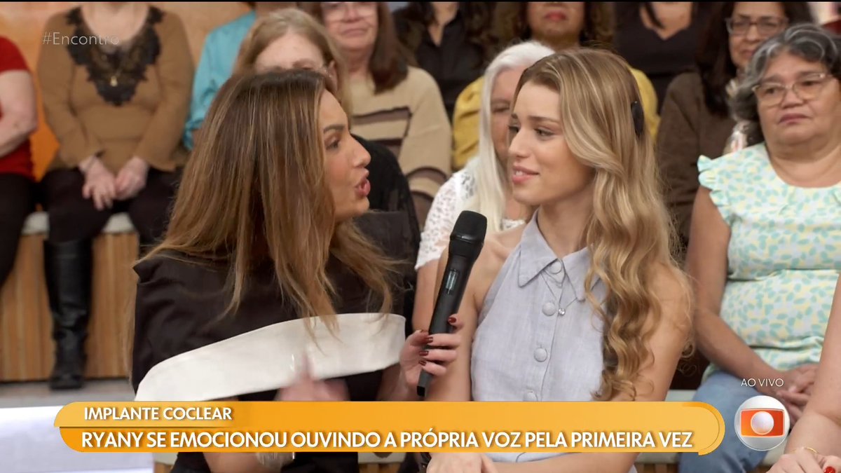 A TV interview scene showing two women with microphones, with a Portuguese caption reading 'IMPLANTE COCLEAR - RYANY SE EMOCIONOU OUVINDO A PRÓPRIA VOZ PELA PRIMEIRA VEZ' alongside an 'AO VIVO' indicator.