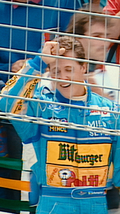 A young Michael Schumacher celebrates with a raised fist behind a metal fence, wearing his iconic Benetton racing suit adorned with sponsor logos including "Bitburger," "Ford," "Suga," "Minol," and "Mild Seven" — capturing the triumphant early moment in the career of the future seven-time World Champion during the 1994 season.