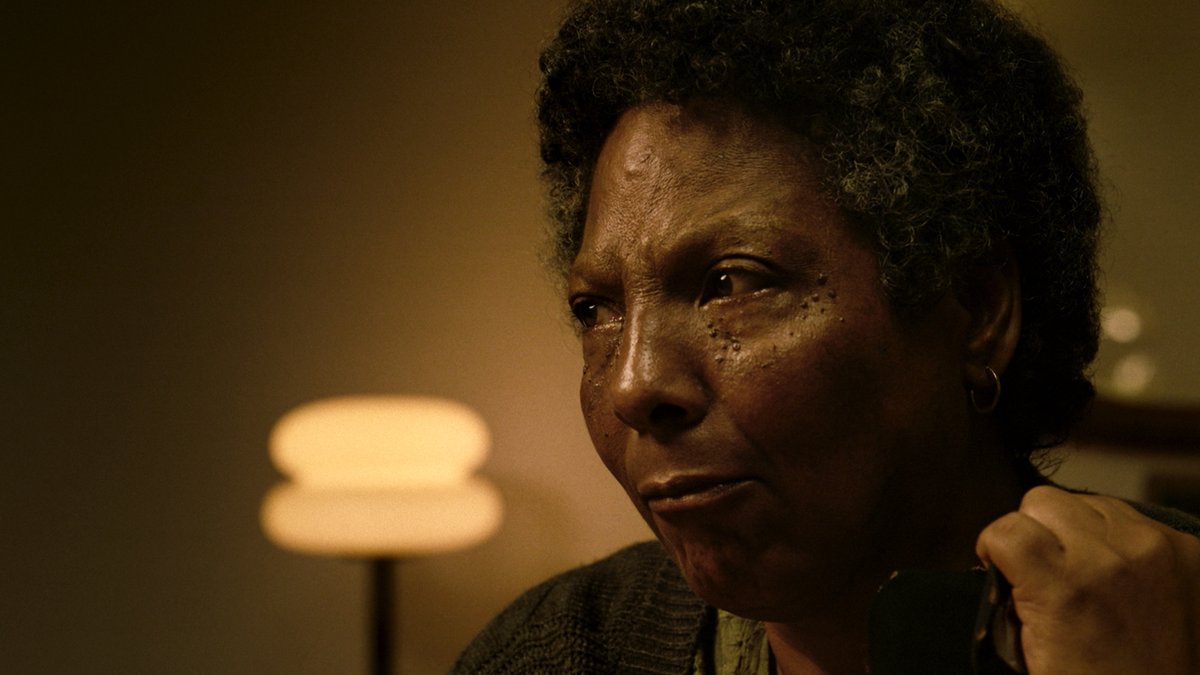 An older woman with short, curly graying hair looks off to the side with a contemplative, tearful expression in dim, warm lighting, her hand resting near her face. The somber, intimate atmosphere of the shot reflects the emotional weight suggested by the post, which promotes the acclaimed Brazilian film *Vicentina Pede Desculpas* and its themes that promise a moving, tear-inducing viewing experience.