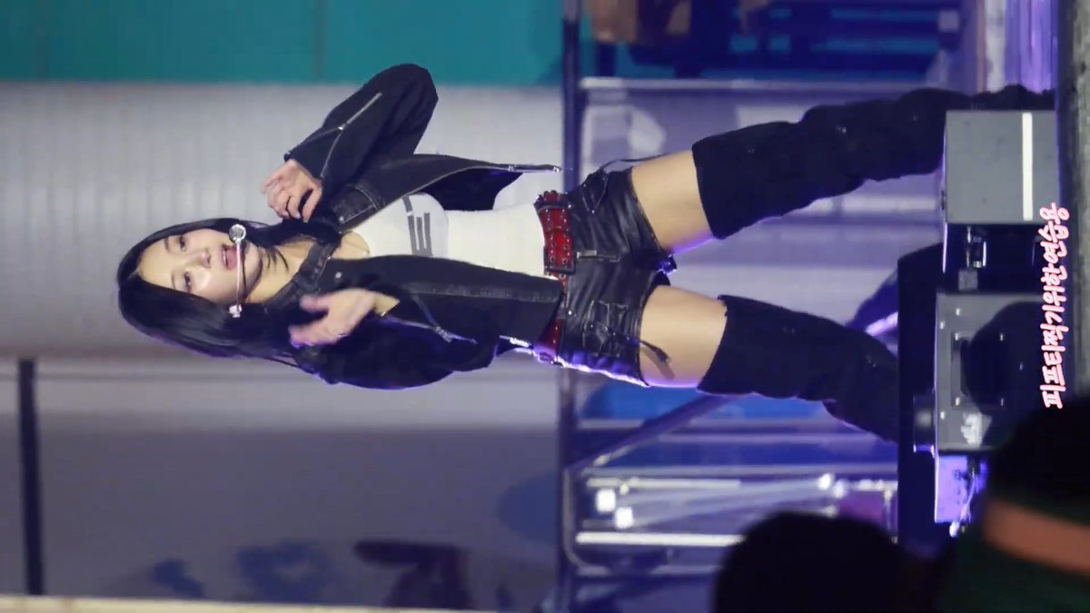 A young woman with long dark hair performs on stage, wearing a black leather jacket, white crop top, and black shorts.